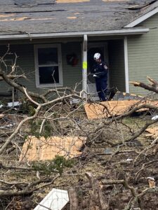 Man standing on front pourch of house that has been damaged and has fallen trees in the front yard